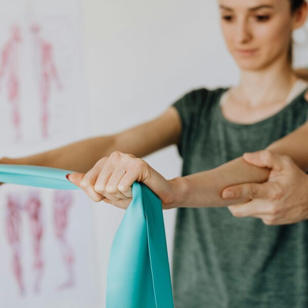 Crop anonymous orthopedist in wristwatch and uniform helping young woman in casual wear reaching arms with elastic tape in doctor office